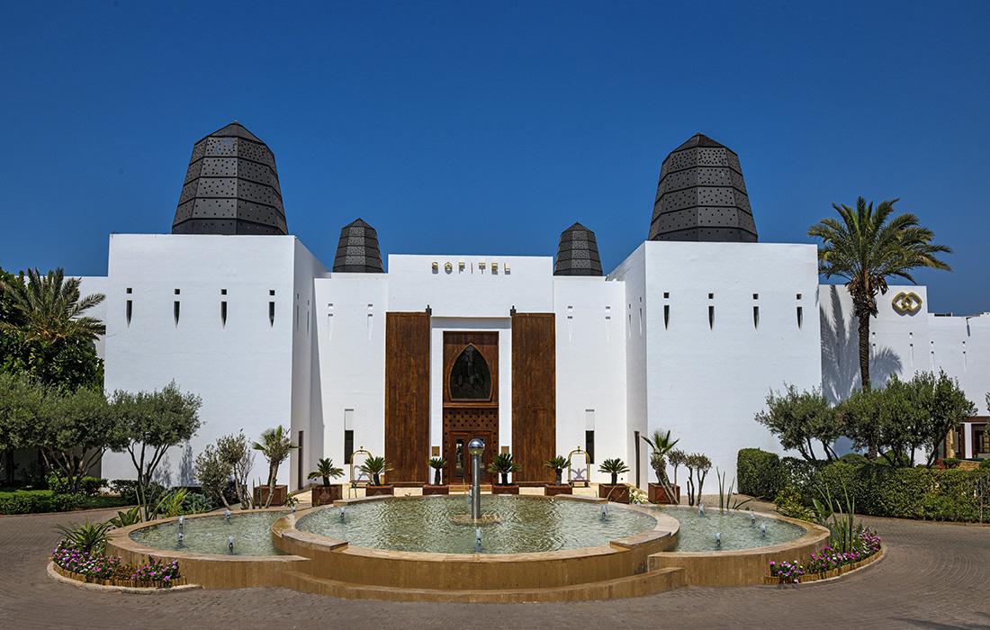 White building with dark conical roofs labeled Sofitel. Foreground features a circular fountain, plants, and palm trees under a clear blue sky. This sofitel hotel in agadir morocco.