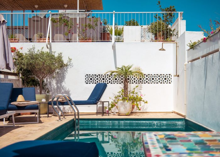 A serene poolside with blue loungers, potted plants, and white walls under a clear sky. A colorful table edge is visible in the foreground.