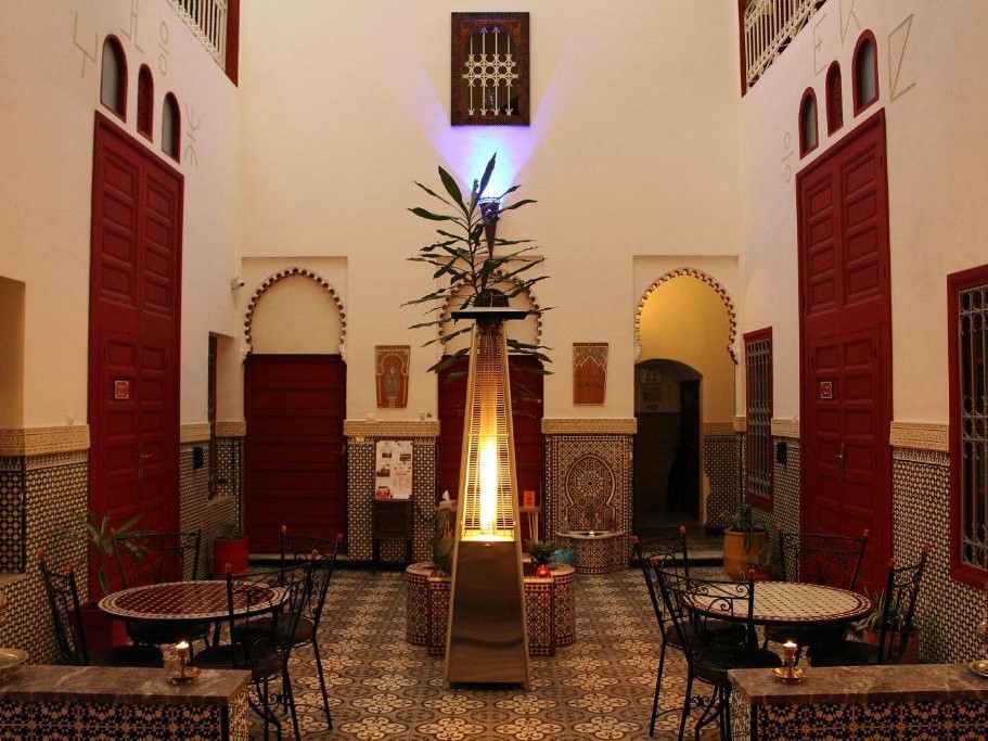 Elegant courtyard with round tables, chairs, and a central tall heater. Red doors and ornate patterns on walls create a warm ambiance.