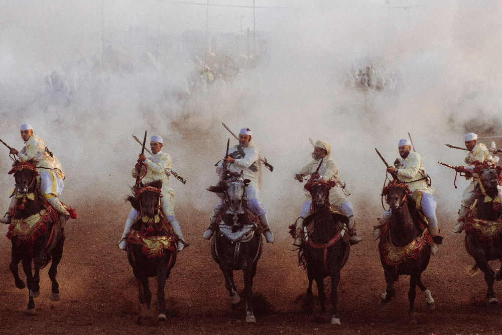 Riders in traditional attire charge forward on decorated horses through smoke, dust rising from a dry, open field, creating a dramatic scene.
