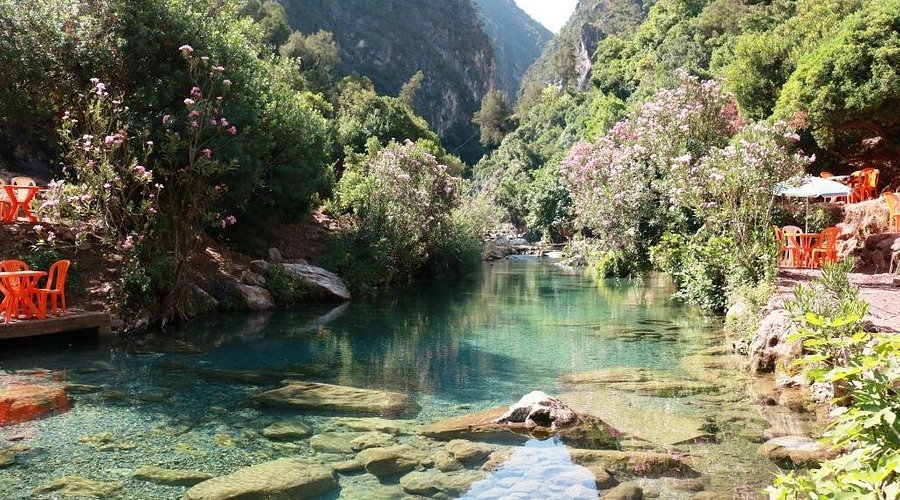 Clear river in a lush green valley with pink flowering bushes. Orange chairs and tables on the bank. Bright, serene atmosphere.