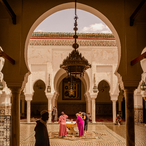People in colorful attire gather around a fountain inside an ornate courtyard with arches, columns, and a large chandelier overhead in fes morocco.