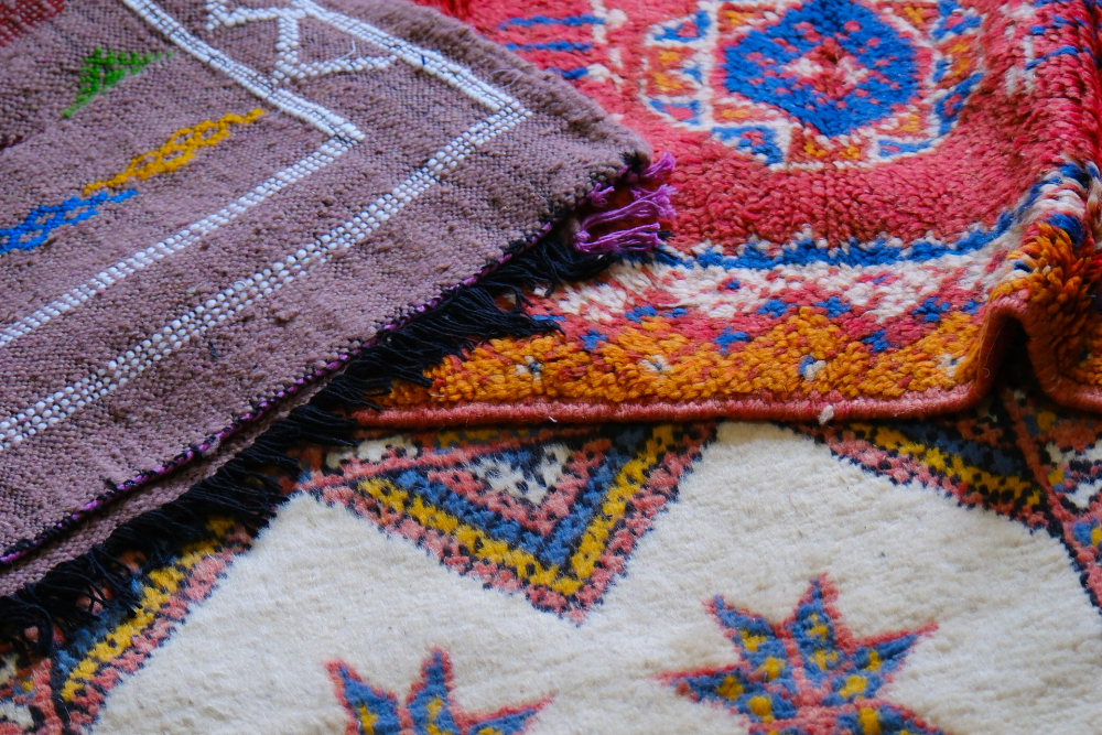 Overlap of three colorful woven Moroccan rugs with geometric and star patterns in red, blue, yellow, and purple, creating a vibrant, cozy mood.