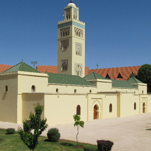 A mosque with green-tiled roofs and a tall minaret stands under a clear blue sky, surrounded by a plaza with small shrubs and trees.