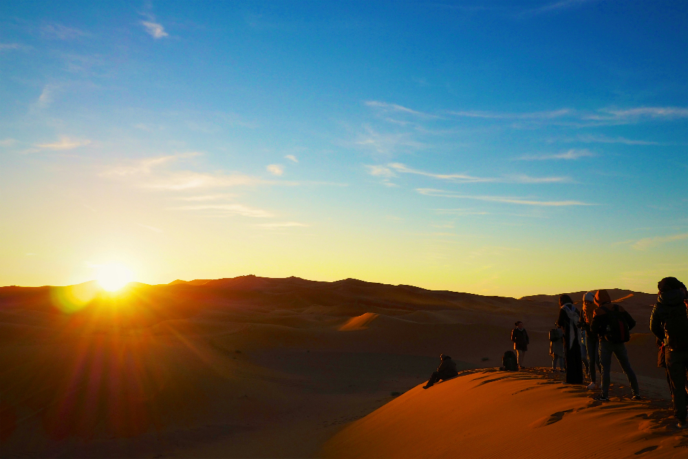 People standing on sand dunes at sunset, casting long shadows. The sky is clear with a warm gradient from yellow to blue. Tranquil scene in erg chigaga morocco.