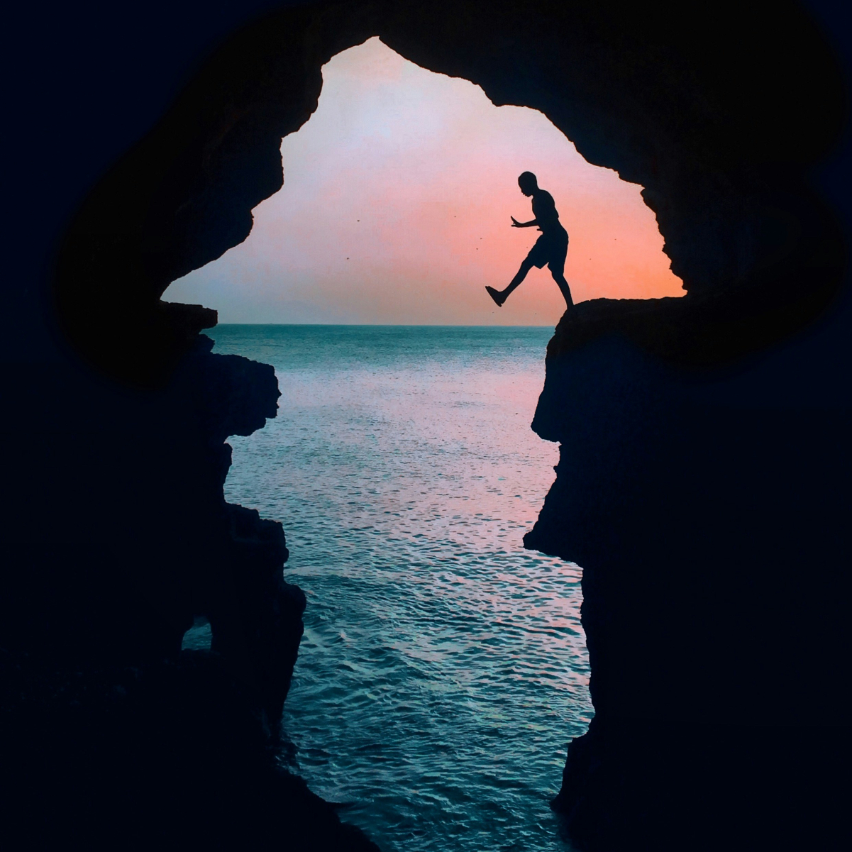 Silhouette of a person jumping between rock formations at hercules caves in tangier morocco at sunset by the sea. Sky is pink and orange, creating a serene backdrop.