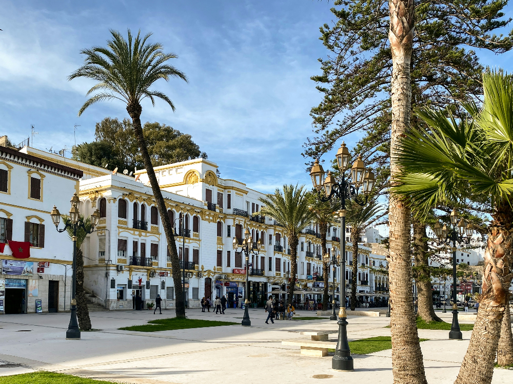 Historic square with white and yellow buildings, palm trees, and vintage street lamps. Blue sky, people walking, and a clear day in tangier morocco.