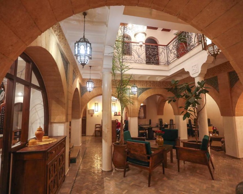 Elegant Moroccan-style interior with arches and lanterns. Green chairs around wooden tables, potted plants, and intricate railings create a cozy ambiance.