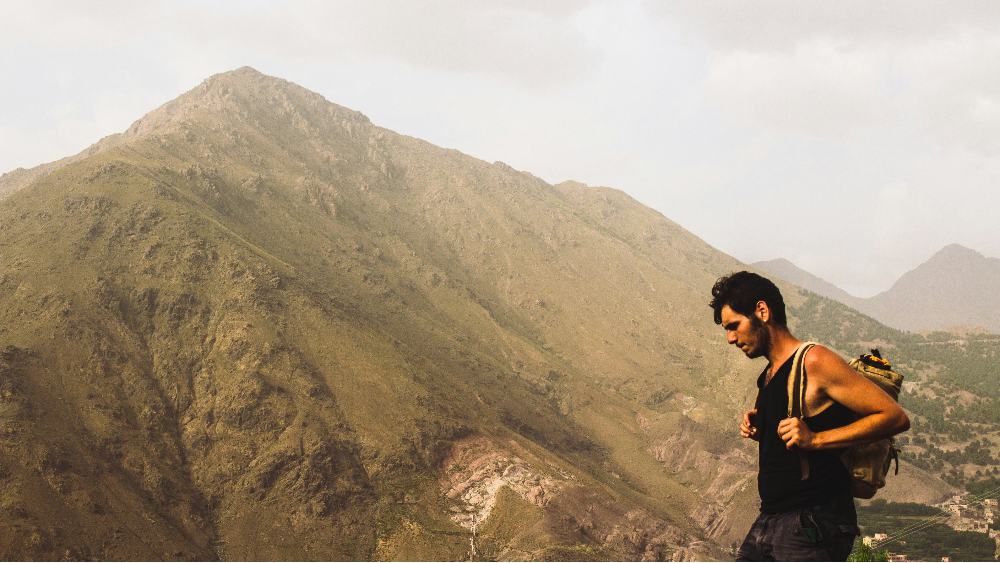 Man with a backpack hikes near a large, brown mountain under a cloudy sky, conveying adventure and tranquility.