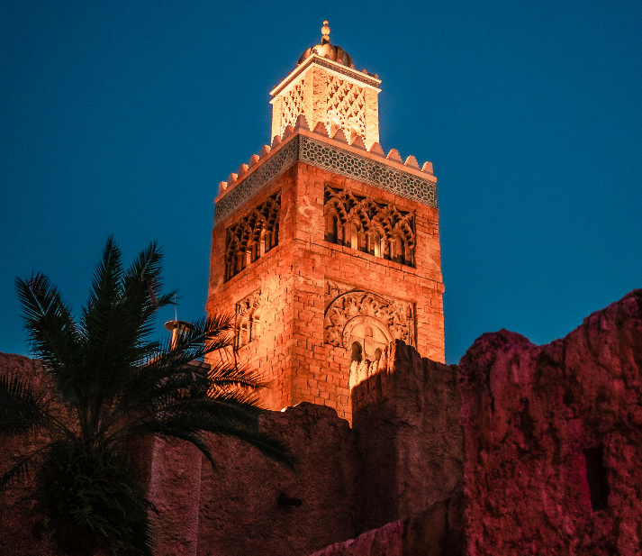 Tall illuminated mosque in Ramadan with ornate patterns against a deep blue sky. Palm tree and stone walls in foreground. Warm, serene ambiance.