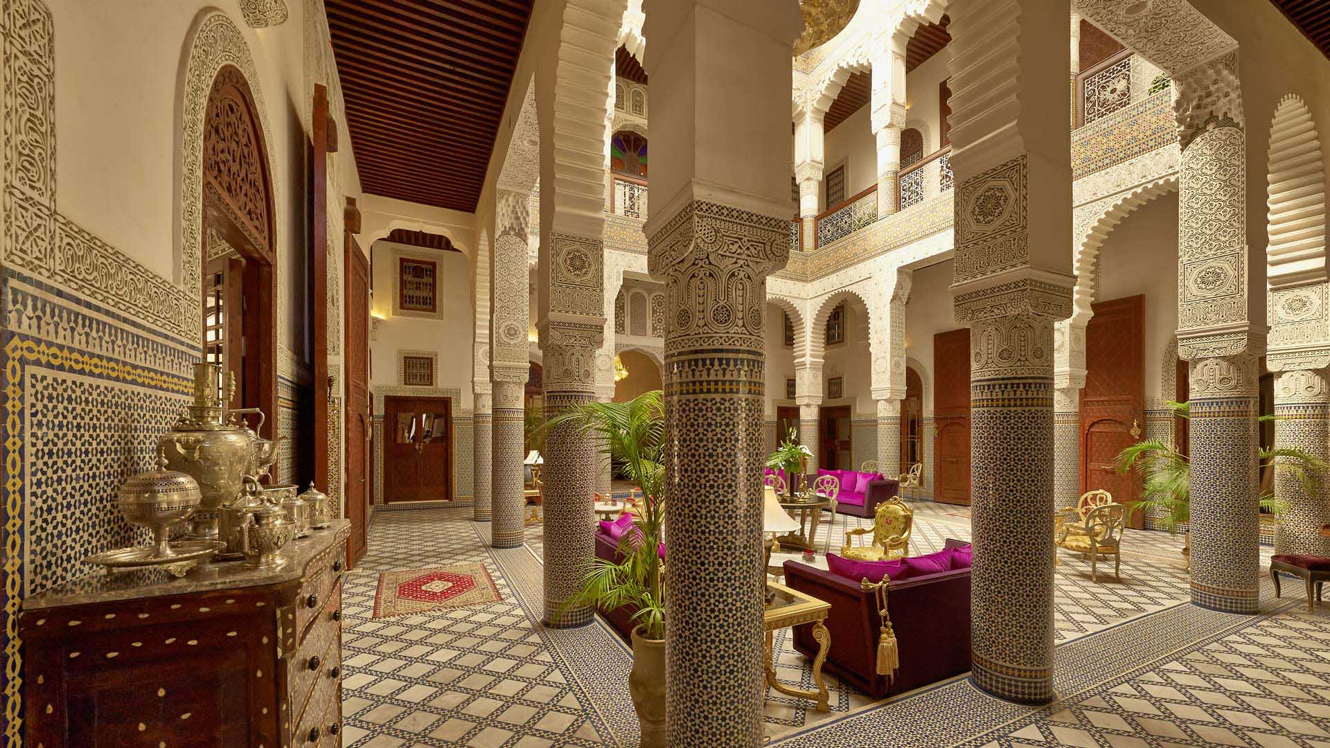 Elegant Moroccan courtyard with ornate columns and intricate tilework. Purple sofas and chairs, silver tea set, and lush plants create a luxurious vibe.