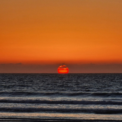 Sunset over ocean with vivid orange sky, red sun at horizon, and dark, rippling waves creating a calm, serene atmosphere.
