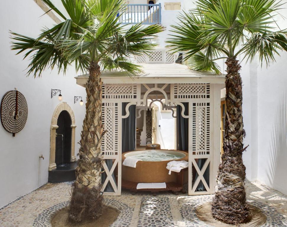 Two palm trees flank a jacuzzi under a decorative gazebo in a sunlit courtyard. The walls are white with a circular patterned art piece.