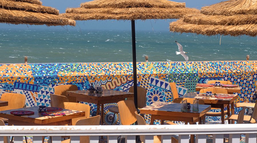 Seaside café with wooden tables and chairs under straw umbrellas. Colorful mosaic wall, ocean waves, and a flying seagull in the background.