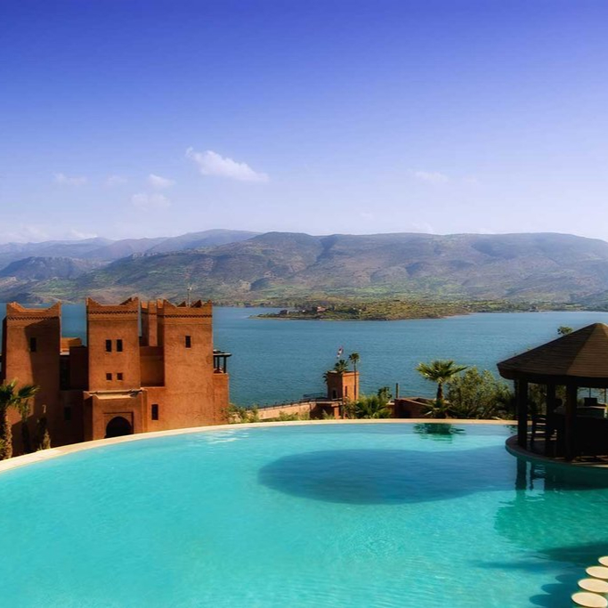 A serene infinity pool overlooks a lake and mountains, with a castle-like brown building on the left under a clear blue sky.