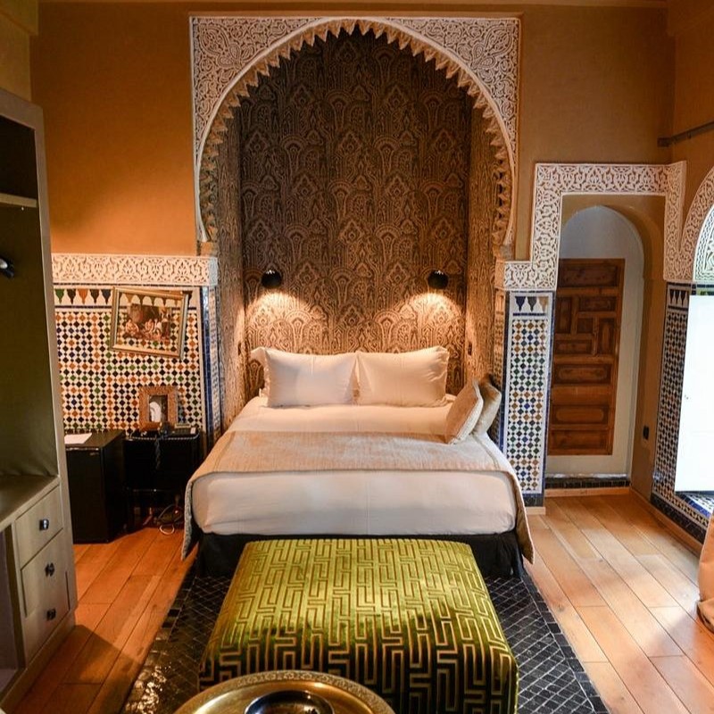 Elegant bedroom with an ornate arch, patterned walls, and a cozy bed. Green ottoman in foreground, warm lighting creating a cozy mood.
