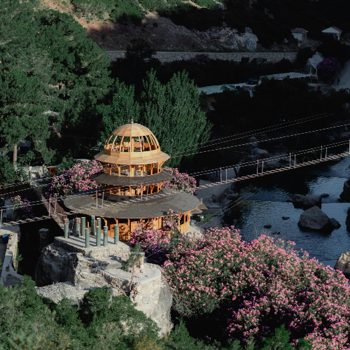 A wooden pavilion surrounded by pink flowers, near a tranquil river and a suspension bridge, set against a lush green hillside.