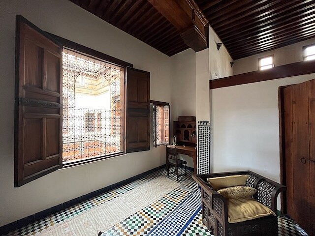 Cozy room with wooden ceiling, open window, patterned floor, and a wicker chair. Light streams in, highlighting the serene atmosphere.