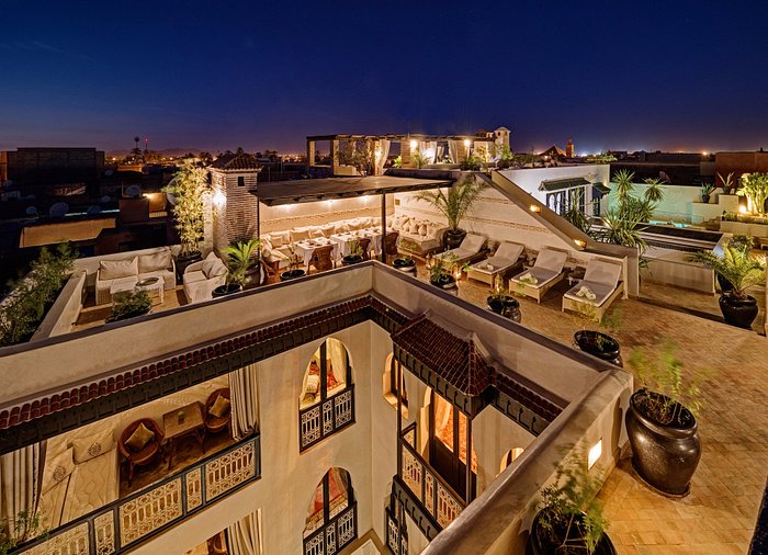 Rooftop terrace at night with lounge chairs, dining area, and lush plants. Warm lighting contrasts with starry sky, creating a cozy ambiance.