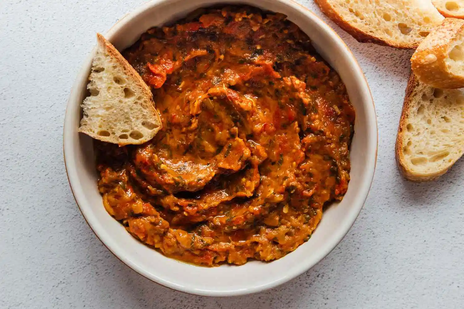 Bowl of zaalouk, which is a Moroccan red-orange vegetable dip with spices, garnished with crusty bread slices, on a light textured surface.