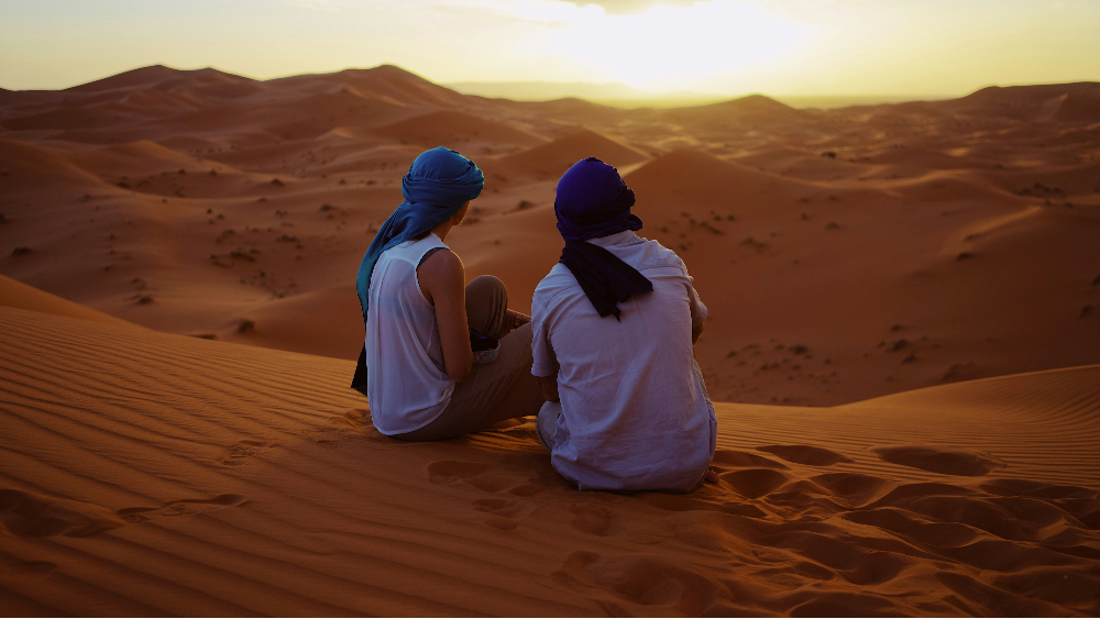 Two people in blue headscarves sit on desert dunes at sunset, gazing at the vast landscape. The sand glows orange, creating a serene mood.