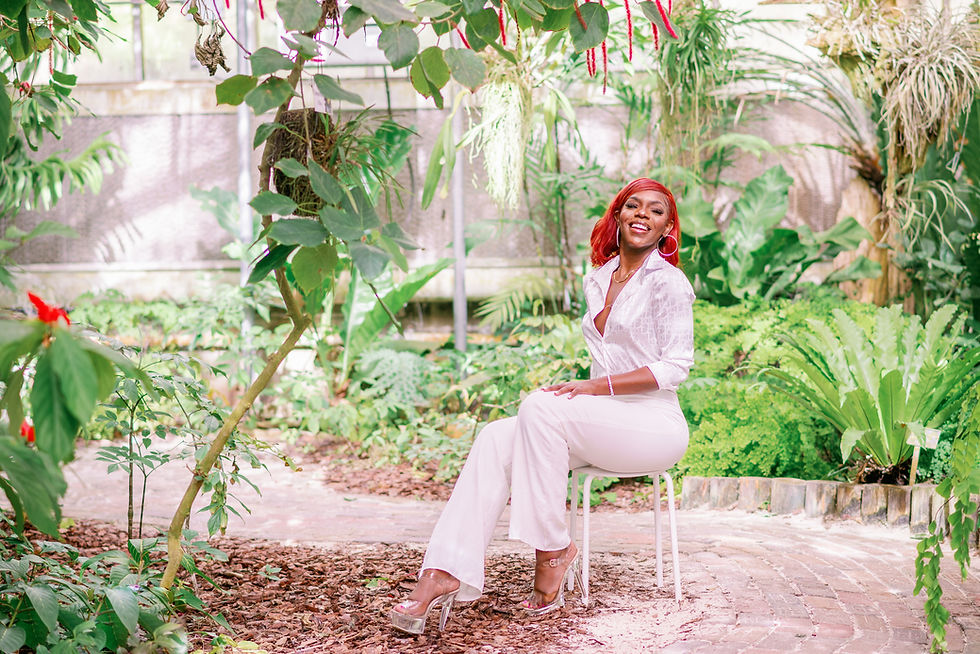 Smiling woman with red hair sits on a stool in a lush, green garden. She wears a white outfit and heels. Sunlight filters through plants. Photo by Tampa headshot photographer Nina Bashaw.