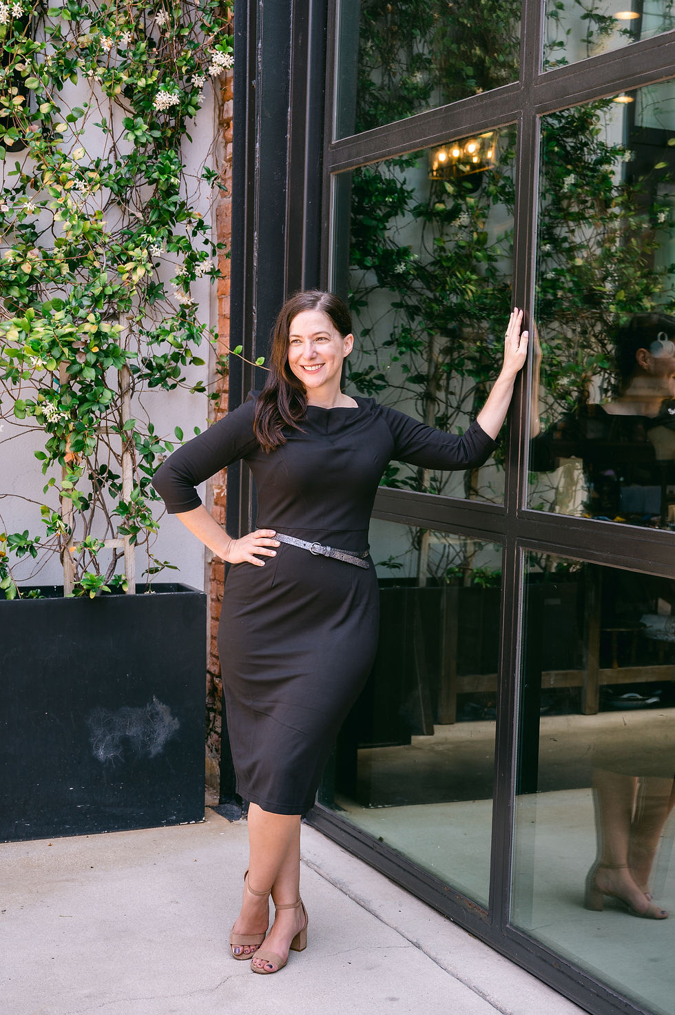 Woman in a black dress poses confidently by a glass door, smiling. Background features green vines on a wall. Mood is cheerful. 10 Outfit Ideas for Women to Elevate Their Headshots. Advice from Tampa Headshot Photographer Nina Bashaw