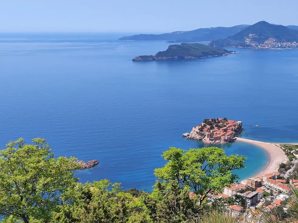 Aerial view of a tiny red-roofed island village, blue bay, curved beach, and distant mountains on a sunny day.