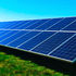 Solar panels in rows on a grassy field under a clear blue sky, with highways in the background. Bright sunlight highlights the panels.