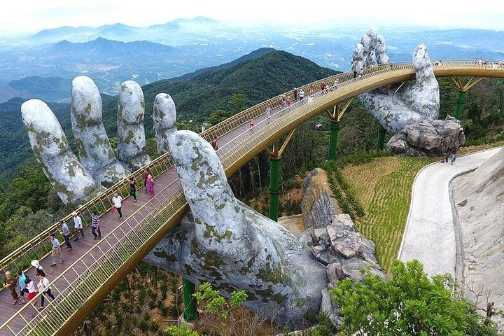 Tourists walk across Vietnam’s Golden Bridge held by giant stone hands, with lush mountains and a misty valley below.