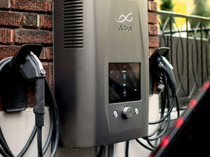 Wall-mounted electric vehicle charger with cables on a brick wall. Display screen active. Mood is functional and modern. VEHICLE T GRID