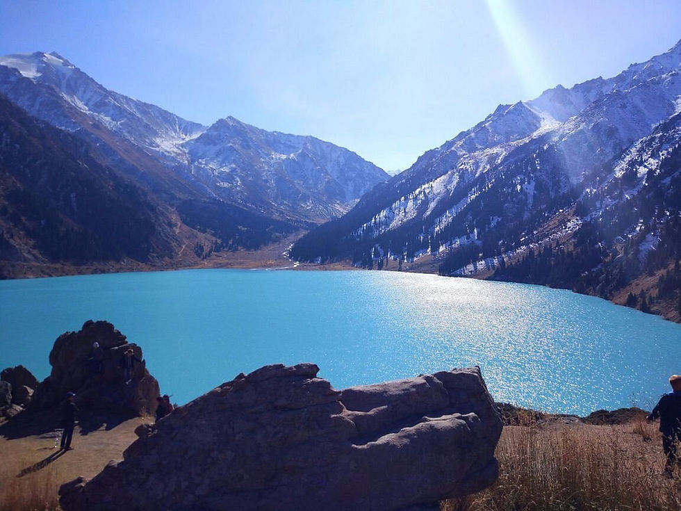 Turquoise mountain lake in a snowy alpine valley, with a few hikers on rocky shore under a bright blue sky.