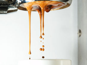 Espresso pours in smooth streams from a metallic machine into a white cup, against a light background. The coffee is rich and creamy.