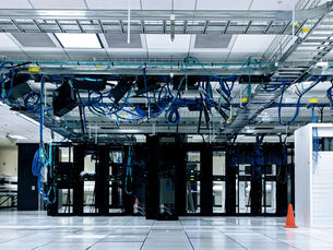 Server room with rows of black and white servers, blue cables overhead. A table and chair are visible nearby, with bright lighting.