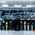 Server room with rows of black and white servers, blue cables overhead. A table and chair are visible nearby, with bright lighting.
