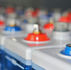 Close-up of a car battery with blue and red terminals, metallic bolts, and a blurred industrial background.