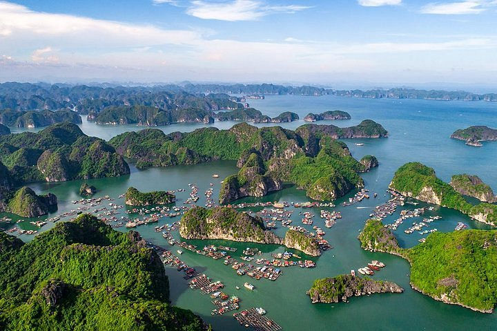 Aerial view of green limestone islands and crowded boats in a blue bay under a partly cloudy sky.