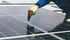 Worker in blue overalls and green gloves uses a drill on a rooftop solar panel, showing focus and precision. Reflective surface.