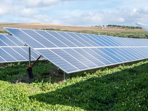 Rows of solar panels in a green field under a blue sky with clouds. Rolling hills in the background. Bright and sunny setting.