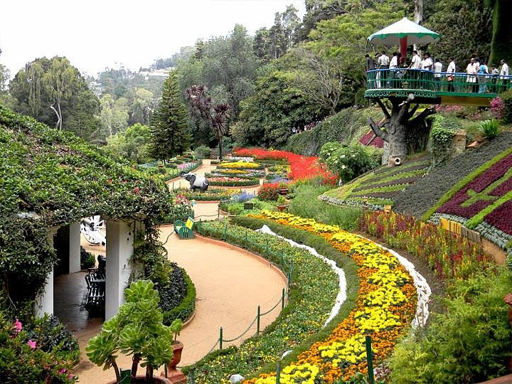 Colorful terraced flower garden with winding path and visitors on a treehouse-style gazebo amid lush greenery.