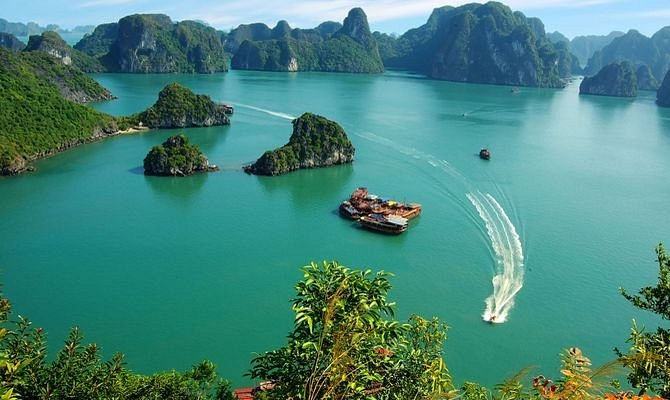 Turquoise bay with limestone islands, boats, and a speedboat leaving a white wake beneath a clear blue sky. Vietnam