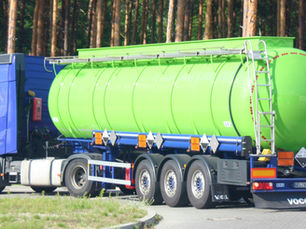 A blue truck with a green hydrogen cylindrical tanker is driving past a forest. The tanker has a ladder, and the truck is on a paved road.