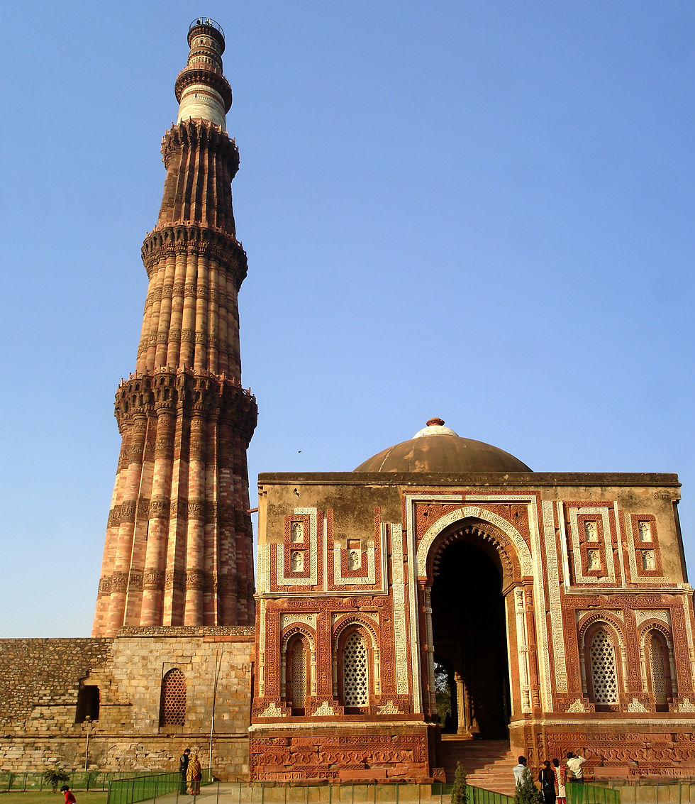 Qutub Minar rises beside an ornate domed mosque under a clear blue sky, with a few visitors below.