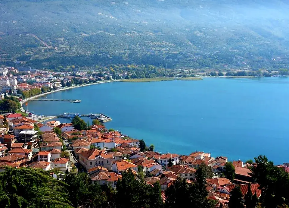 Aerial view of a lakeside town with red-tiled roofs, a blue bay, a small pier, and green mountains under clear skies