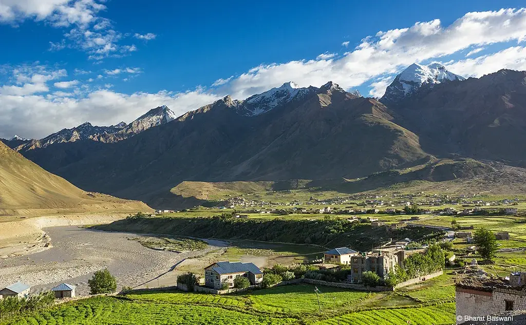 Sunlit mountain valley with river, green fields and village below snowcapped peaks under a blue sky; watermark reads Bharat Baswani.