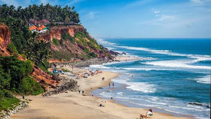 Cliffside path above a sandy Varkala beach and calm blue ocean, with green plants and waves below under a clear sky.