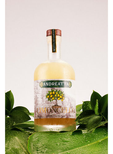 Miniatura: Foto frontal do Limoncello Andreatta com aspecto gelado em um ambiente temático da natureza, folhas verdes rodeiam a garrafa