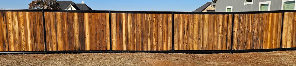 Wooden fence with vertical planks and black trim in a yard. Grass and dirt in foreground, houses in the background, clear blue sky.