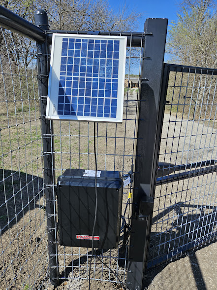 Solar panel and control box on a metal gate in a rural setting. Sunny day with trees in the background. Blue and black tones dominate.