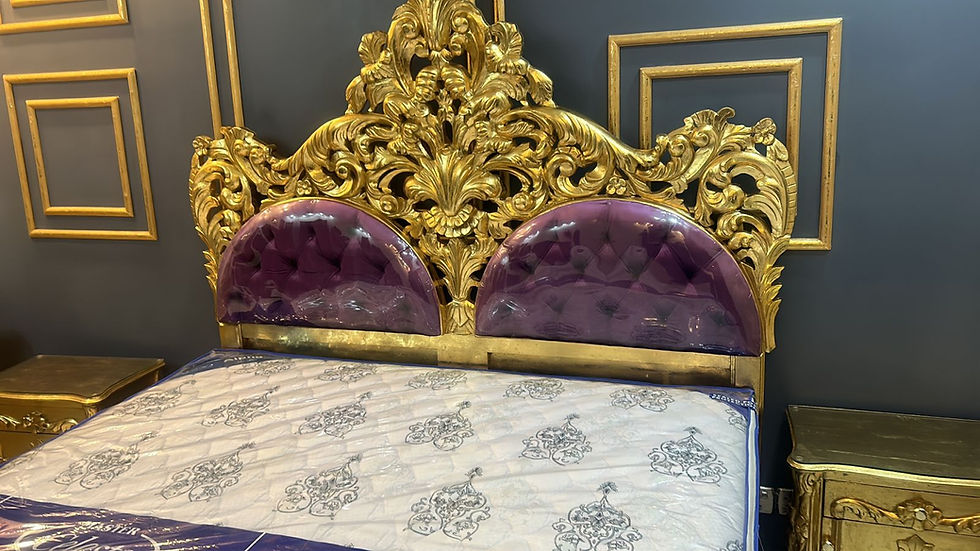 Ornate gold bed with a lavish purple headboard in a room with a dark wall and gold accents. White mattress features elegant patterns.